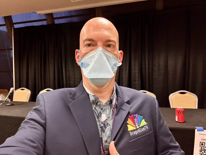 Selfie of a bald masked person wearing a DragonconTV blazer in a convention panel room.