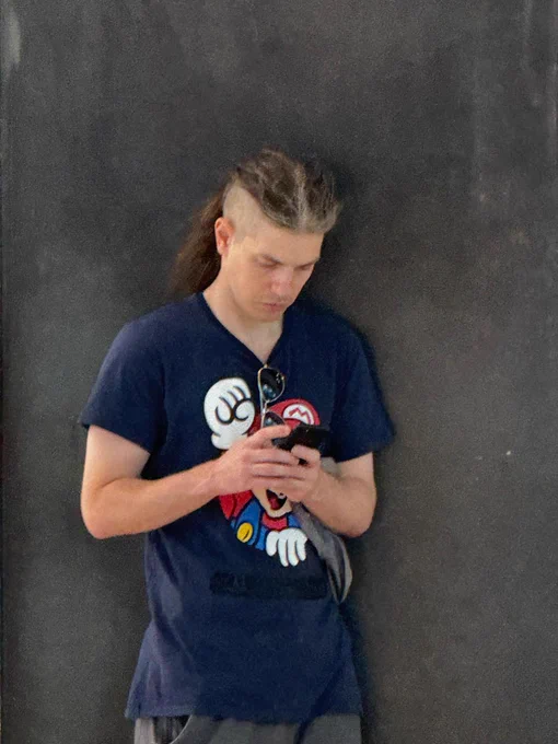 A man with a ponytail and an undercut is looking down at his phone and texting in front of a grey background.