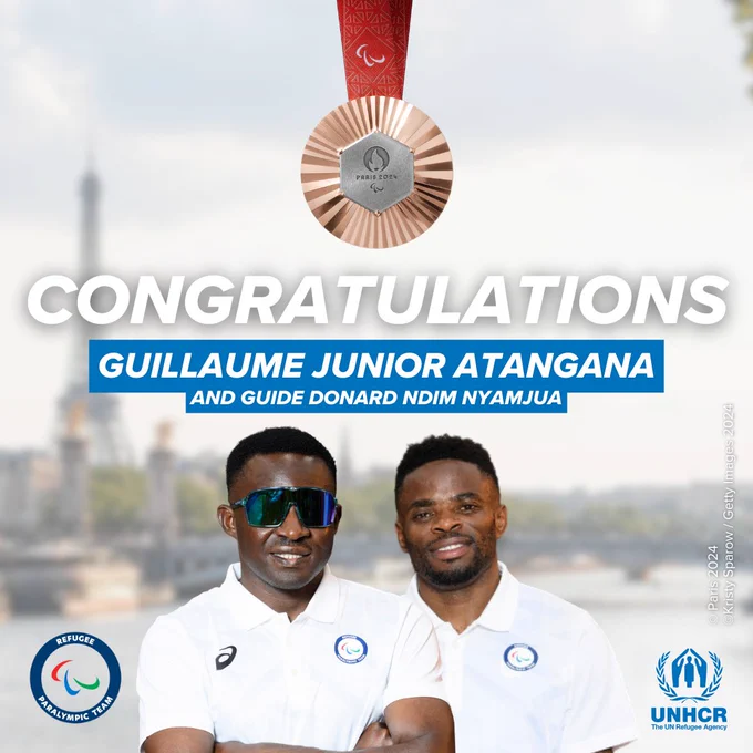 A man stands with his arms crossed, wearing a white t-shirt with the logo of the Refugee Paralympic Team on it and colourful sunglasses. Next to him is a man in the same white t-shirt with the Refugee Paralympic Team logo. Above is a bronze medal with the inscription ‘Paris 2024’, and at the bottom of the visual logos for Paris 2024 and UNHCR. The background features a blurred cityscape with the Eiffel Tower. The text reads ‘CONGRATULATIONS GUILLAUME JUNIOR ATANGANA AND GUIDE DONARD NDIM NYAMJUA’ in bold blue letters.