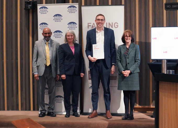Four people stand in front of a banner, showing their logo of the Australian Academy of Science. One of the people holds a certificate.. 