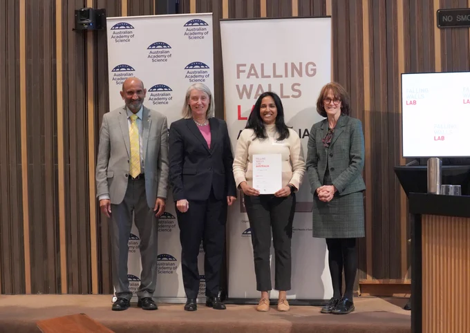 Four people stand in front of a banner, showing their logo of the Australian Academy of Science. One of the people holds a certificate.. 