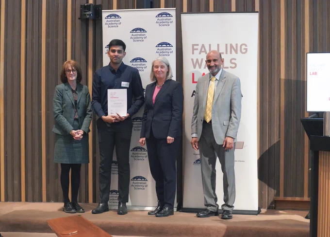 Four people stand in front of a banner, showing their logo of the Australian Academy of Science. One of the people holds a certificate.. 