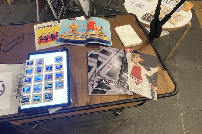 写真。机のうえにマウントされたポジフィルムや冊子や本や雑誌が並んでいる。