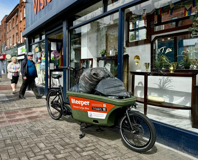 Foreground: a green cargobike with 5 large bags in the front cargo box. 

Background: A sunny day. Window of a charity shop with lots of household items. A man and woman entering.