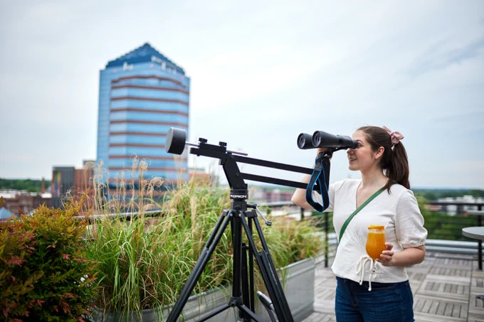 A person holds a cocktail and looks out a pair of binoculars pointed to the sky. In the background is the Durham city skyline.