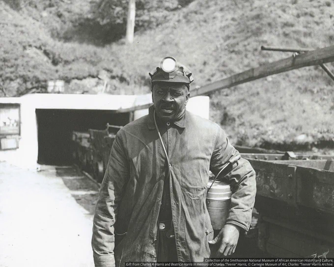 A black and white photograph of a coal miner outside the entrance to a mine. He stands in the center of the foreground; he wears a helmet with an electric light affixed to the crown. He holds a metal lunch pail to his side under his proper left arm. To his right is a line of mine carts parked on a rail leading to the mine entrance. The back of the photograph is blank.