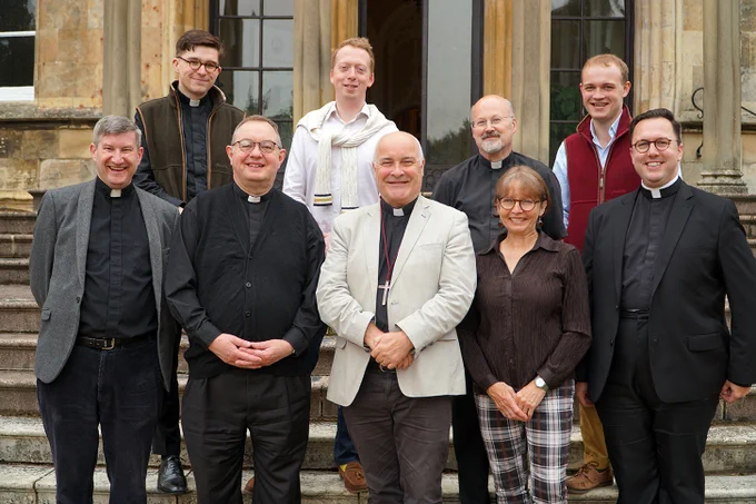 Group of eight smiling clergy members and one layperson standing in front of a historic building.