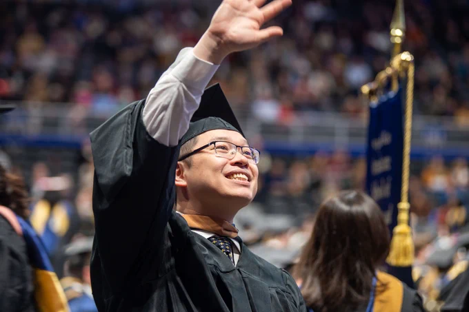 Graduate in cap and gown smiling and reaching up during a graduation ceremony.
