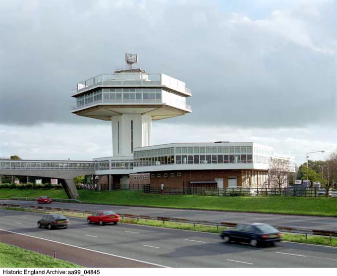 Colour photograph looking across a motorway towards a service station. The service station includes a polygonal section elevated on a brick lower section. To the side is a hexagonal tower with a cantilevered hexagonal structure topped with a sun deck. To the left of the photograph is a covered footbridge spanning the motorway.