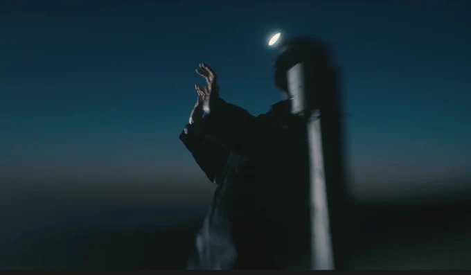 Documentation from a performance taking place outdoors at dusk. A human figure is in the middle of the photo, wearing black loose clothes and a head torch, and raising their arms towards the sky.