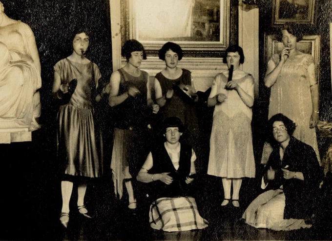 Sepia photograph of a seven female students gathered in front of the fireplace in the Picture Gallery playing instruments. The students are wearing 1920s dresses and have short hairstyles. The instruments being played include cymbals, a tambourine, a Swanee whistle and two kazoos (other instruments are too dark to identify in the image). There are paintings on the wall behind them and a classical style sculpture is visible on the left.