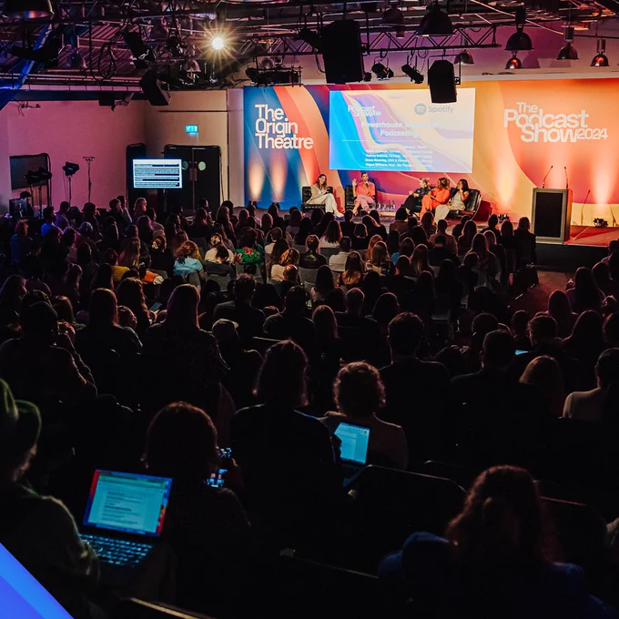 The audience in the Origin Theatre at The Podcast Show 2025 look on as five women speak on stage in a panel. The stage is bright and colourful. Several people in the crowd are taking notes on laptops.