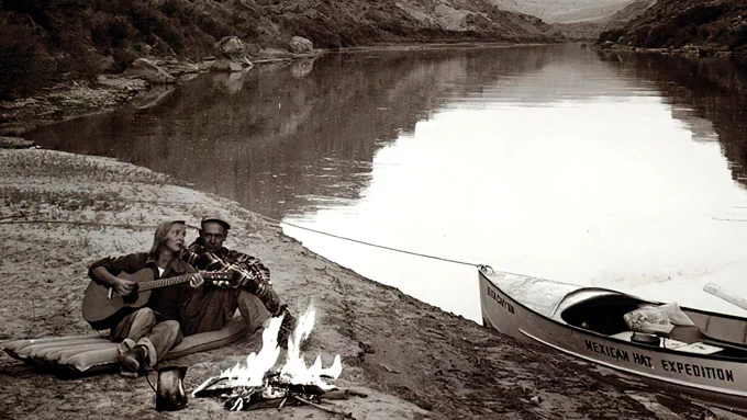 A sepia toned photograph of Katie Lee and Ken Sleight sitting by a campfire near their boat on a river in Utah. The two sit on an air mattress; Katie plays an acoustic guitar. Image by Tad Nichols.