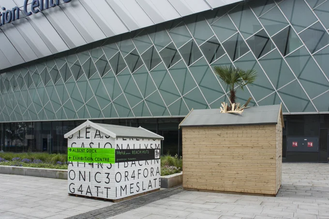 Two beach huts in front of a large glass building. The beach hut on the left is white with black random lettering painted on it, and a bright green banner painted across, with black text which shows the way to the ‘Albert Dock’ and ‘Exhibition Centre’. The beach hut on the right is made up of wooden panels and has a palm tree breaking through the roof of it.