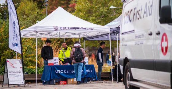 UBC Campus Security tent set up on UBC campus, two people behind the booth and one visitor to the booth talking together. 