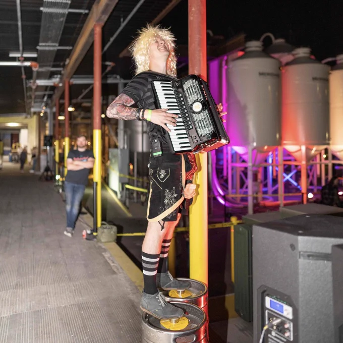 A man in a long hair metal wig stands on two kegs playing the accordion