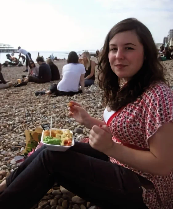 Me, a young white person with wavy dark hair in a deep side parting, sitting on Brighton's pebble beach, balancing a polystyrene container of fish & chips & mushy peas on my knees. Chip in one hand, I'm smiling slightly, but also giving off the vibe that I'd like the person taking the pic to stop photographing me so I can get back to my food. 

I'm wearing a red, black, and white abstract-patterned empire-waist top with black skinny jeans. Behind me is the shell of the old burned-out pier, and the beach is full of people in tshirts & jeans/trackies eating & throwing stones into the sea. 

It feels like the first weekend that the weather has been warm enough to sit on the beach, so everyone's enthusiastically flocked there, but it's still a little chilly if we're honest.