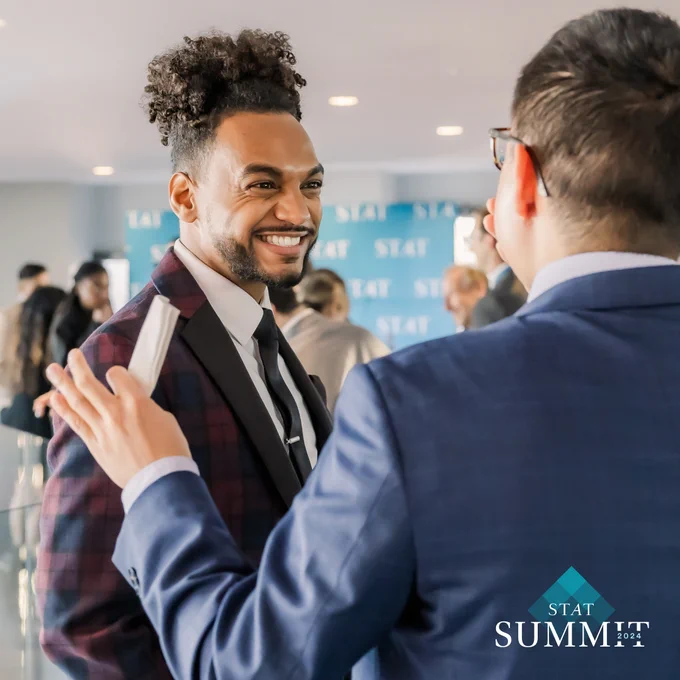 A photo of STAT's Nicholas St. Fleur dressed in a suit and tie as he smiles and embraces another person dressed in a suit with their back turned. The STAT Summit watermark is in the bottom right corner of the photo.