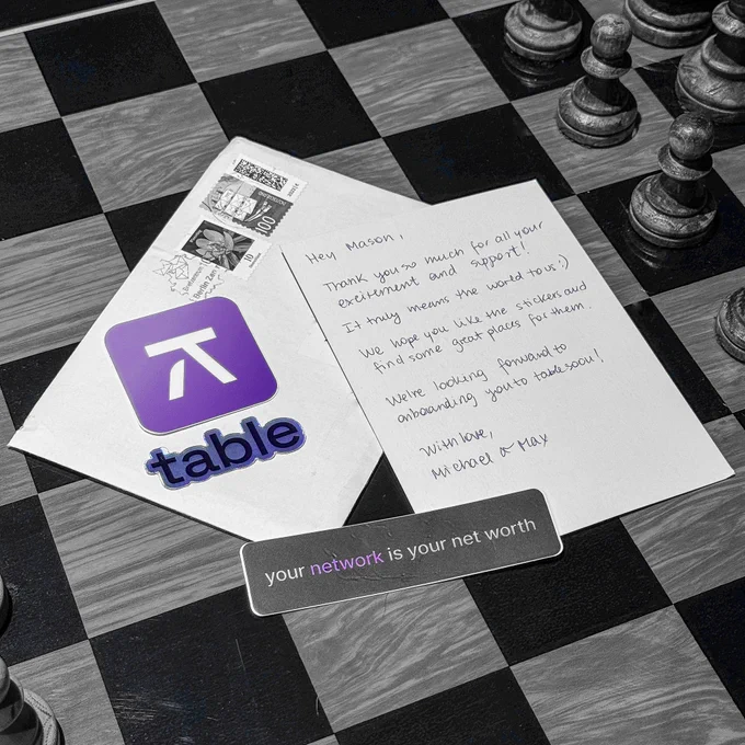 A black-and-white photo with selective color highlighting: a postcard, envelope, and stickers featuring the Table logo are arranged on a chessboard. The purple color is selectively shown in the Table logo, the word “Table” on one sticker, and the word “Network” on another sticker, with the rest of the image in black and white.