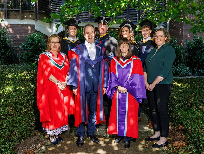 A group of four students standing in a row at a graduation ceremony. In front of them is four lecturers, smiling at the camera.