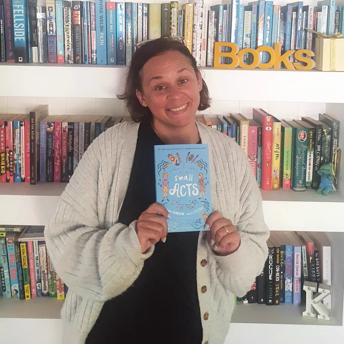 A smiling woman standing in front of a bookshelf and holding a copy of Small Acts