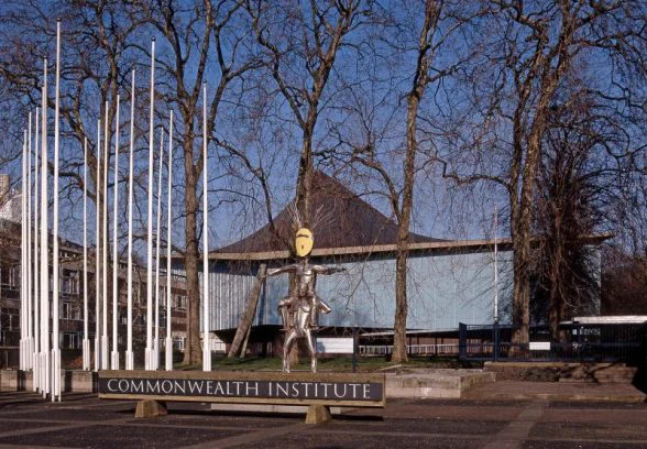 A view of the Commonwealth Institute with a cluster of flagpoles on the left and a silver metal figurative sculpture of two figures in the foreground. One figure has a yellow mask and sits on the shoulders of the other.