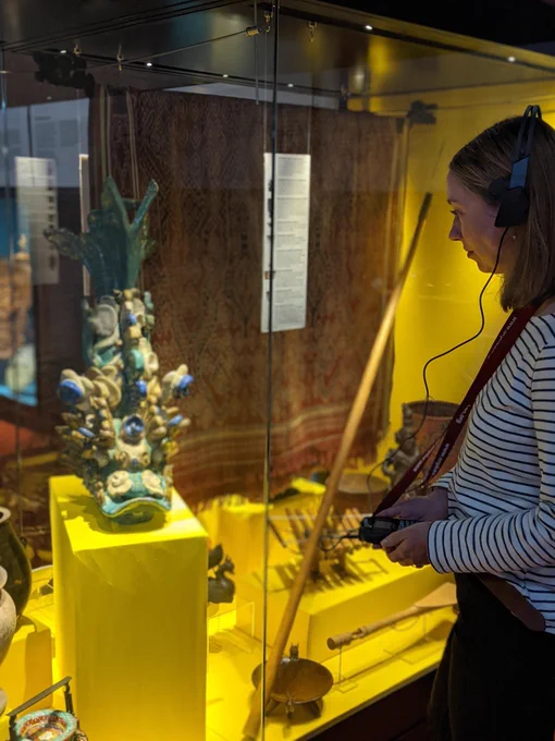 Colour photograph of a person looking at a museum  objects in a glass case while wearing a headset and holding a handset.