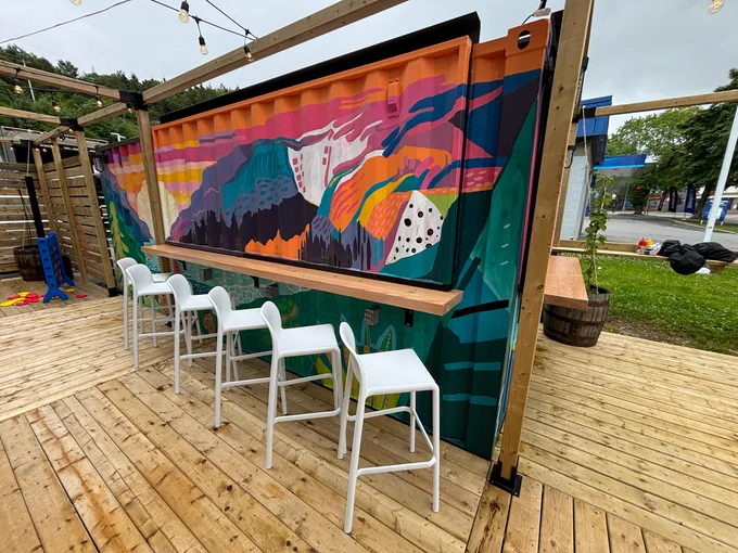 Outdoor seating area with white stools in front of a colorful abstract mural, set on a wooden deck with string lights above.