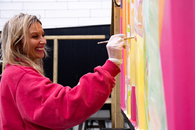 A woman with long, blonde hair, wearing a pink sweater, smiles as she holds a paintbrush up to a colourful wall.