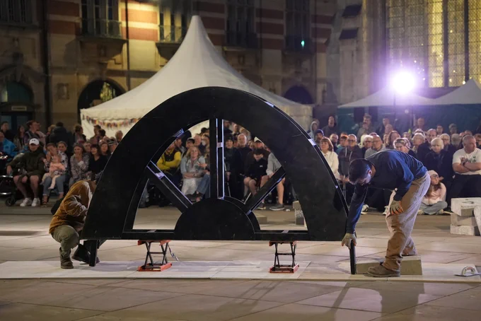 Outdoors at night in an open city square, Kaleider's performance installation Arch begins. Two performers are setting up a wooden former in the shape of an arch. Behind them, rows and rows of audience sit and stand, watching.