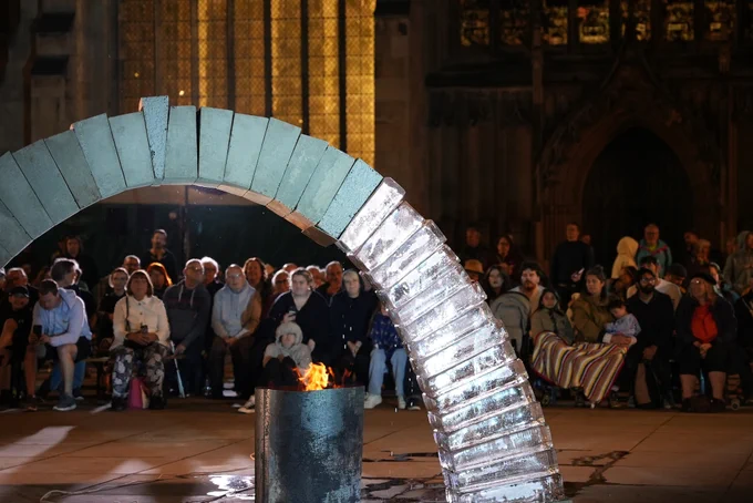 A freestanding arch made two thirds of concrete and one third of ice stands in the square. A fire burns beneath it, melting the ice, as dozens of people wrapped in blankets look on.
