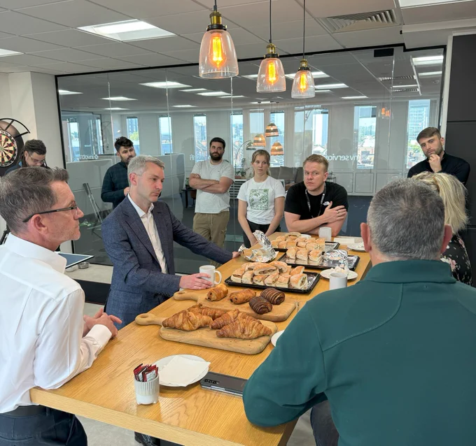 Representatives from Inv Group and its subsidiary companies are gathered in the kitchen area of the Inv Group offices, sharing breakfast and chatting to Will Forster, MP for Woking. In the centre of the table they are standing around is a selection of breakfast pastries and behind the people the offices can be seen through a glass divide.