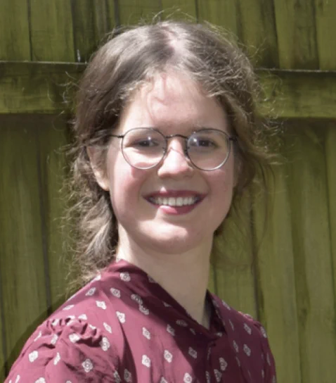 A wood fence in the background. A women in red shirt with brown hair and round glasses.