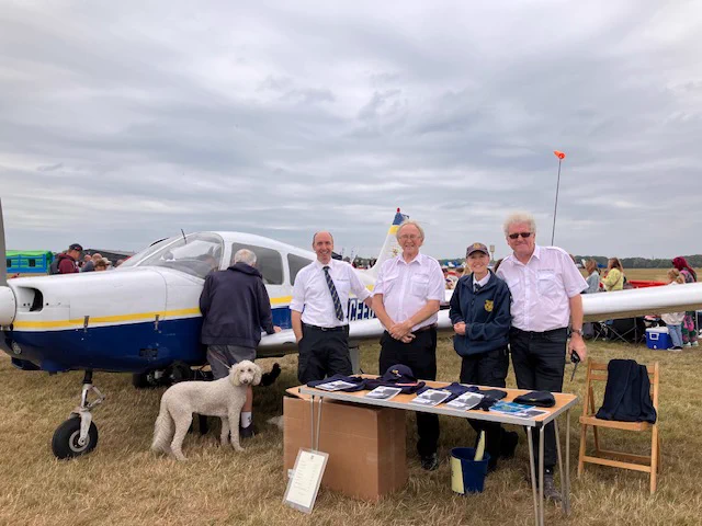 Image of a group of three men and one woman stood by a stall in a busy field. A small white and blue biplane is parked behind them.