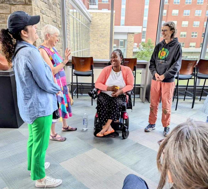 Four women are in a circle: three are standing, one is in an electric wheelchair. All of the women are laughing and one holds her hand up to express something she has just said. These women are in front of a large window facing the central campus residence hall at UVM.