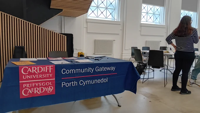 A desk to the left covered with a blue banner with Cardiff University's logo and Community Gateway text, translated also to Welsh. On it, literature and clipboards. To the right, a table with chairs and people standing by it, including a woman with long brown hair in a black and white striped shirt seen from behind. The room is white and lofty with radiators on wall segments underneath windows with a lead "x" effect. To the left, behind the table, an angled staircase with vertical wood slats making up the railing.