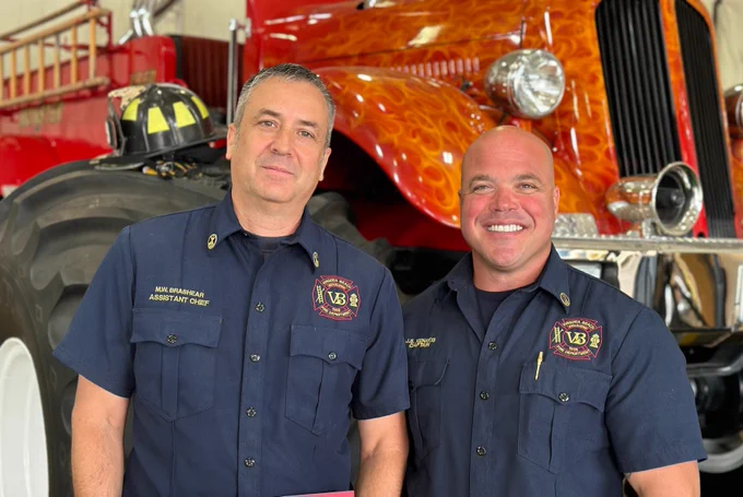 Two firefighters in uniform smiling in front of a monster fire truck.