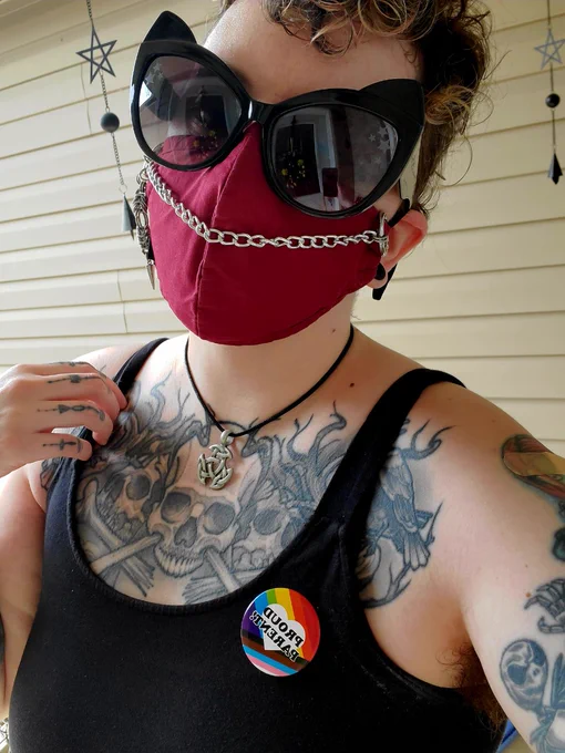 A White Non-Binary person with various tattoos on arms, chest & hand,  wearing big black cat eyed sunglasses, a deep red n95 face mask with a silver chain on it, on their neck is a silver Celtic knot on a black necklace chain and they're wearing a black tank top with a Pride Flag pin that reads "Proud Parent" on it.