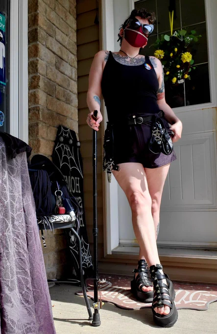 A White Non-Binary person wearing big black cat eyed sunglasses, a deep red n95 face mask with a silver chain on it, black tank top, shorts & sandals holding onto a black medical cane, on a front porch with black Halloween doormat, welcome sign and table cloth seen.