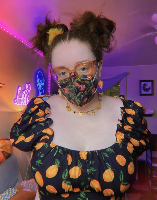 Me, wearing a black lemon top covered in lemons with a frilly neckline, a floral KN95, orange pie earrings, a gold necklace, glasses, and hair in messy high buns, standing in my living room.