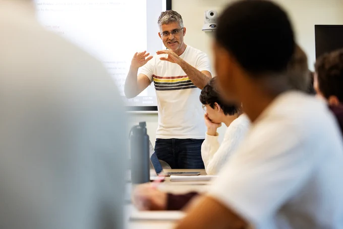 Professor Riera-Crichton is gesturing while lecturing and can be seen two students in the foreground.