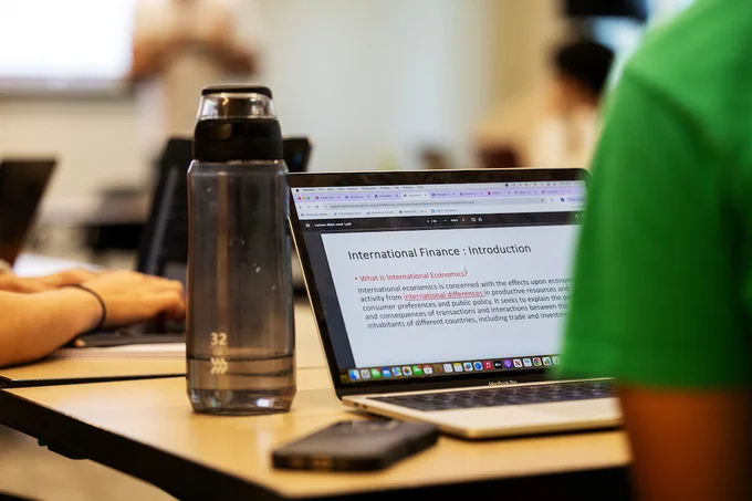 A water bottle (on left) and a student's laptop (on right) can be seen while the student (off screen to the right) takes notes on their laptop during class.