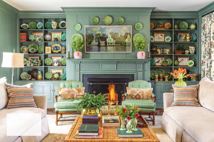 Alt text: A swipe-through image of a cozy living room with shelves and a fireplace painted in a light cool-toned green and adorned with vintage majolica plates, artwork, and antique collectibles. Accenting the room are cozy matching cream sofas and green armchairs. 
