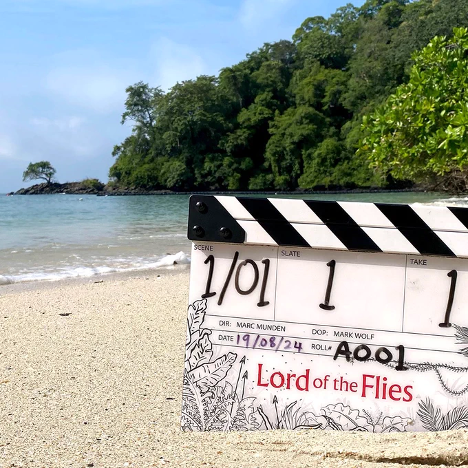 A clapperboard is pictured in sand on a beach. It's illustrated with drawings of plants and speaks and the words Lord of the Flies are printed on it in red. Green trees and vegetation are visible in the background, where the sea gently rolls in to meet the sand.

