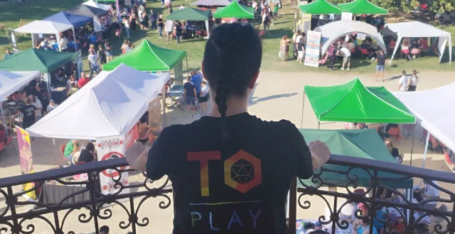 Una persona dello staff di To Play (la manifestazione ludica torinese) osserva da un balcone i numerosi tavoli da gioco, coperti da tende e gazebo e occupati da centinaia di giocatori, che riempiono il Parco della Tesoriera. La fotografia è scattata alle sue spalle, e sul retro della sua maglietta compare il logo dell'evento.