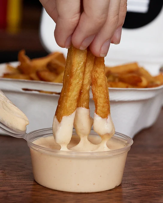 A person dipping 3 fries into a cup of house sauce, with additional fries visible in the background.