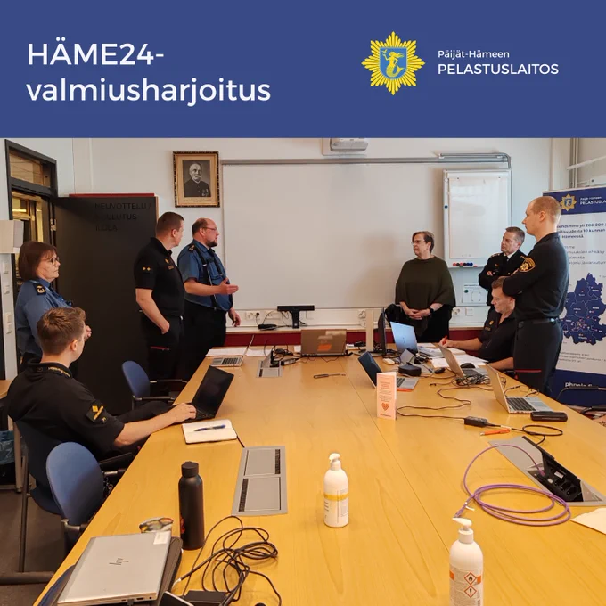 Kuvassa teksti HÄME24-valmiusharjoitus ja Päijät-Hämeen pelastuslaitoksen logo. Kokoushuoneessa otettu kuva. Iso neuvottelupöytä. Yksi siviilivaatteissa oleva henkilö sekä kahdeksan pelastustoimen virkavaatteissa. Kaksi istuu pöydän ääressä tietokoneiden edessä, muut seisovat. 