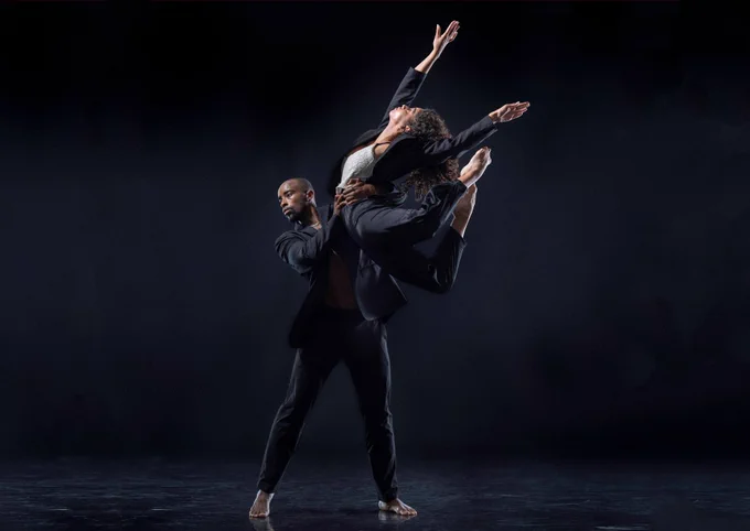 A male dancer lifts a female dancer, both wearing dark clothing, in an expressive pose against a dark background.