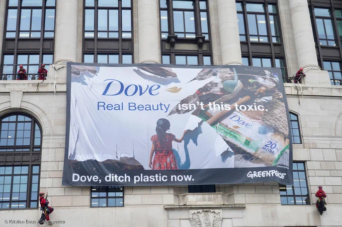 Climber activists unfurl artwork on Unilever's HQ in London. The artwork reads "Dove - real beauty isn't this toxic - dove ditch plastic now. The artwork shows a young child peeling back Dove's branding to expose plastic pollution.
