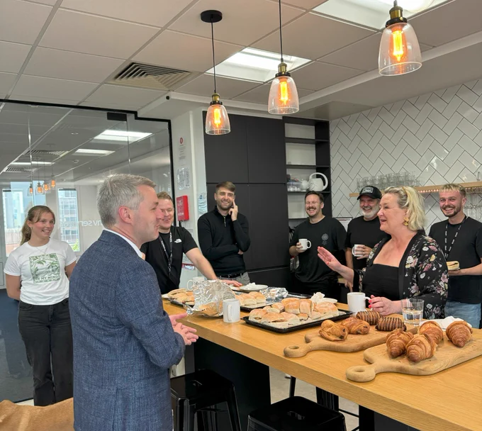 Will Forster, Liberal Democrat MP for Woking and Alison Galvin, MBE are chatting across a table covered in a selection of breakfast pastries. Representatives from Inv Group and its subsidiary companies are gathered around them sharing breakfast and chatting. Behind the people the offices can be seen through a glass divide.